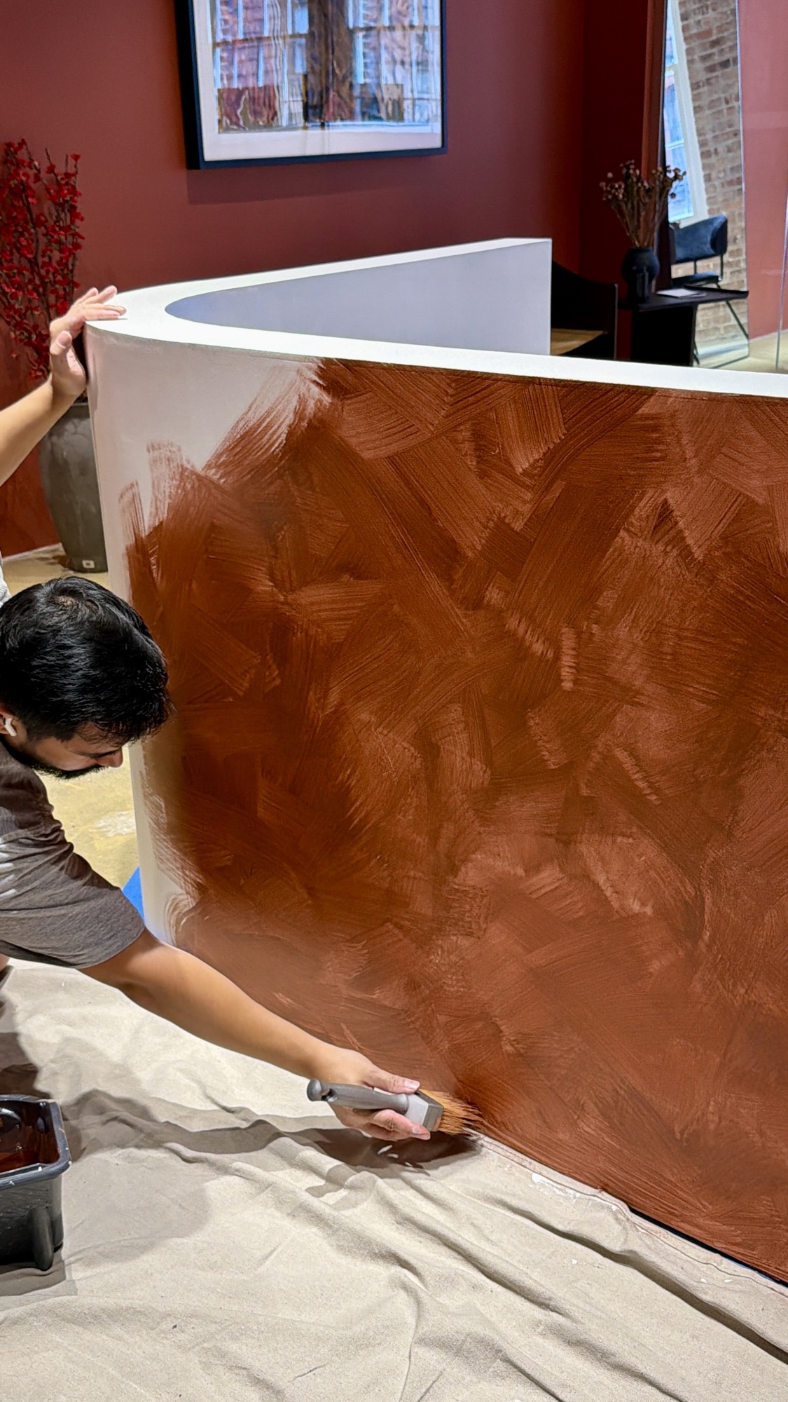A Limewall & Co. painter hand-brushing terracotta limewash onto a curved showroom desk, the cross-hatched brushwork visible